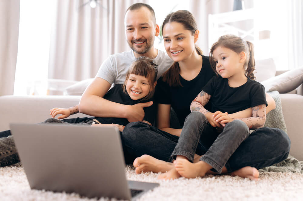 Young Parents and Their Two Amazing Daughters Communicate via Video Conference With Grandparents Sitting at Home on the Floor