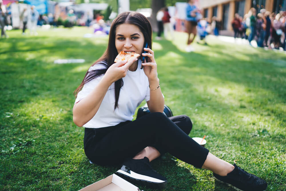 Positive Caucasian Female Resting on Festival in Park Eating Tasty Pizza Talking on Smartphone