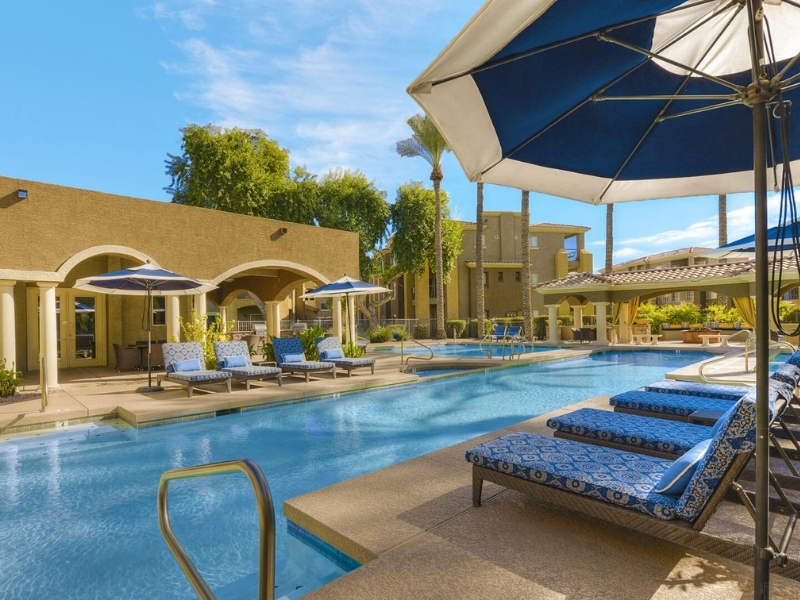 Resort pool area with blue patterned lounge chairs under umbrellas along the water’s edge, sunlit buildings in the background.