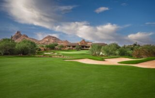 Desert golf course with green fairways, sand bunkers, shrubs, and a distant clubhouse under a blue sky.