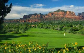 Golf course with green fairway, flag near the hole, yellow wildflowers in the foreground, and red rock mesas in the distance under a blue sky.