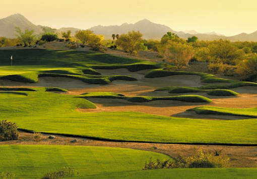 em2-1 Panoramic view of a desert golf course with rolling green fairways, sand bunkers, and distant mountains at sunset.